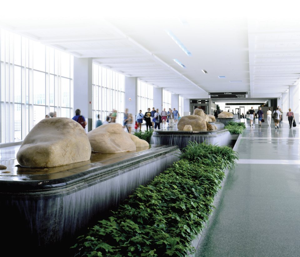 A bright airport walkway with water features, large boulders and a planted strip, with travelers walking toward the security checkpoint