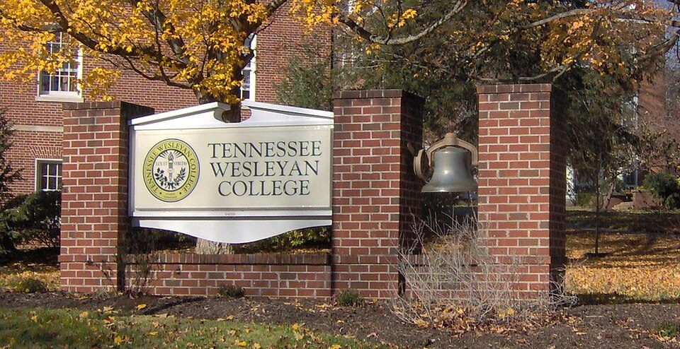Brick campus sign reading Tennessee Wesleyan College beside an old bell, with autumn leaves